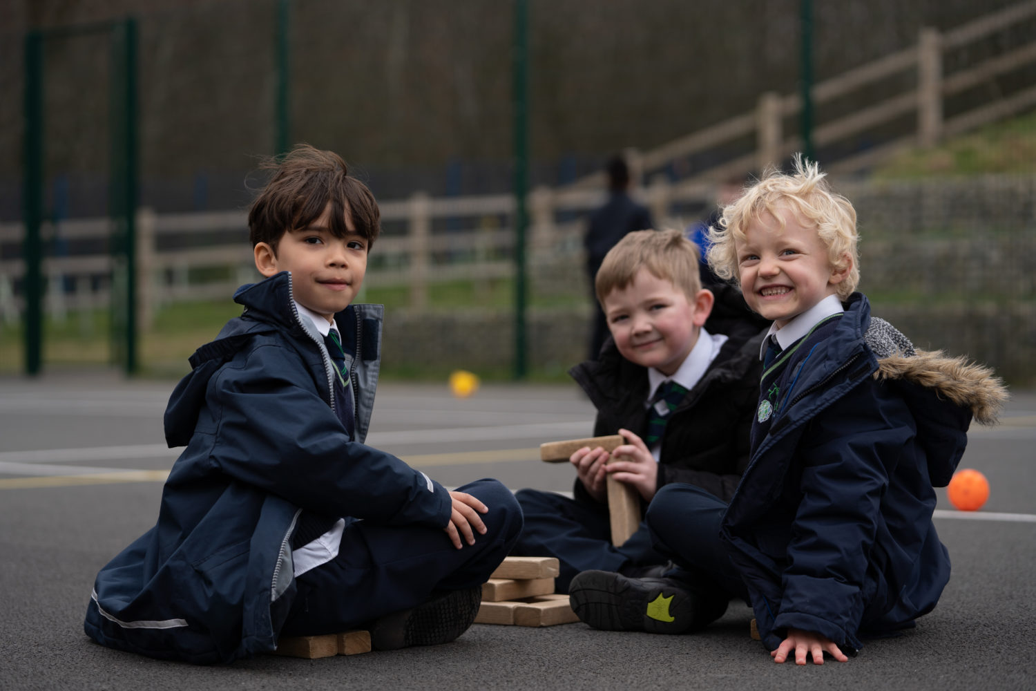 Three young boys are shown sitting on the ground and playing with wooden blocks together in an outdoor area on the academy grounds. All three are wearing their winter coats.