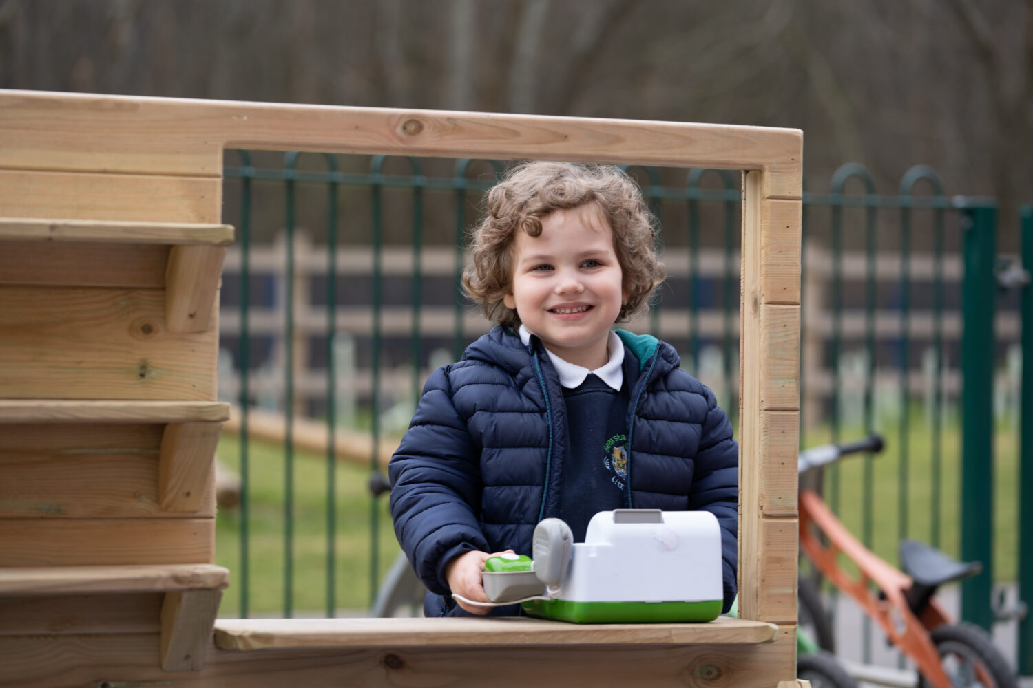 A young boy with long, curly hair is seen smiling through a hole in a wooden fence and wearing his winter coat.