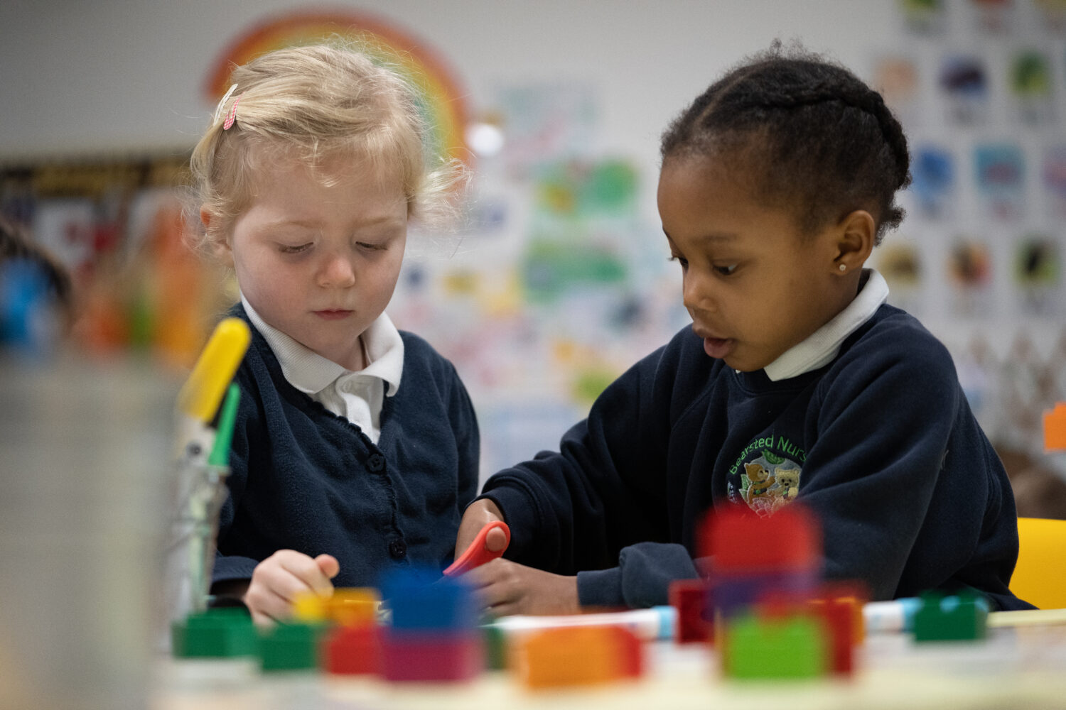 Two young Nursery girls are pictured participating in an Arts & Crafts session together. The girl on the right can be seen cutting through paper with a pair of scissors.