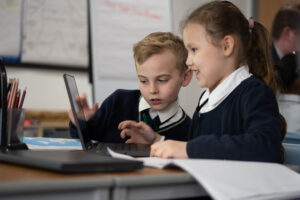 Two young pupils, a boy and a girl, are seen working together on a laptop computer. The girl is seen smiling whilst looking at the screen.