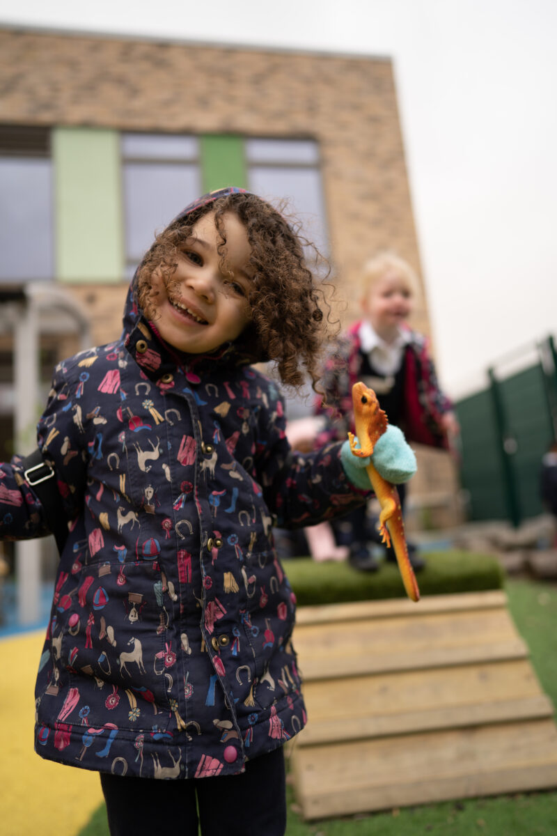 Two young girls are seen playing together outdoors in their winter coats. One of the girls is holding a toy Lizard in her left hand and smiling at the camera.